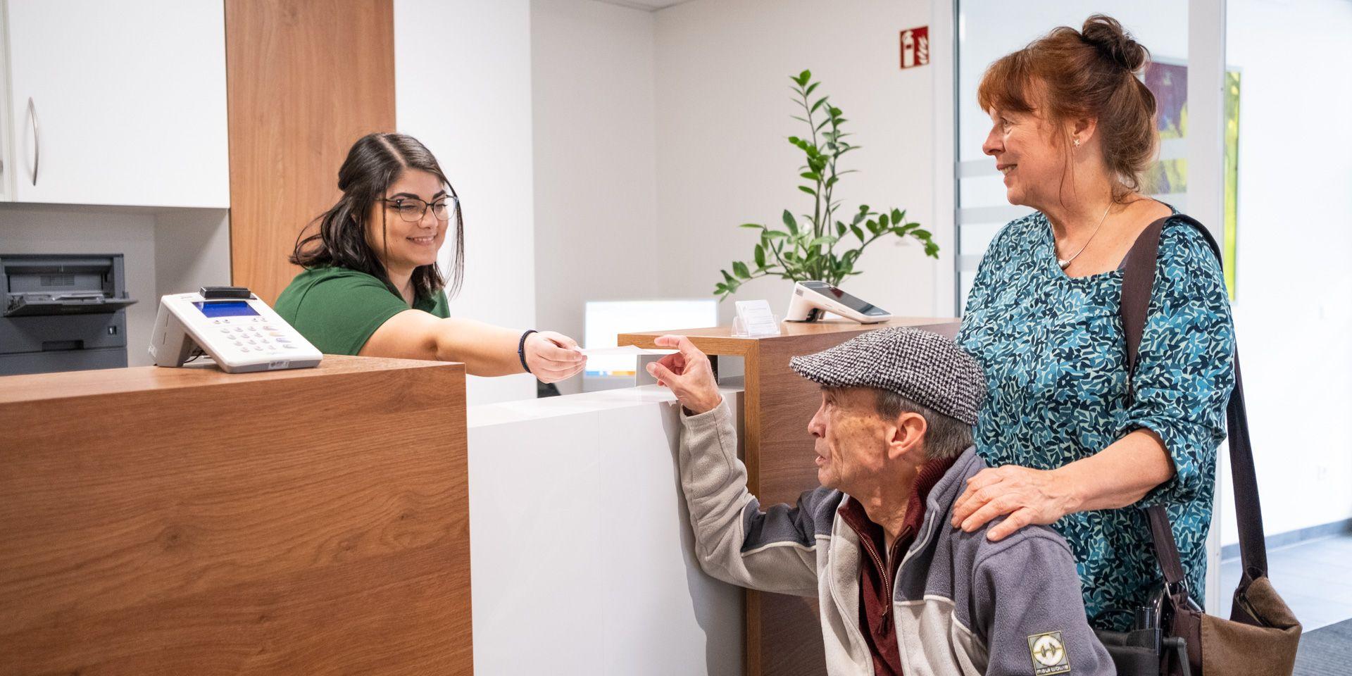 Arzthelferin an der Anmeldung gibt einem Patienten im Rollstuhl seine Versichertenkarte zurück, begleitet von einer Frau. Arzthelferin an der Anmeldung gibt einem Patienten im Rollstuhl seine Versichertenkarte zurück, begleitet von einer Frau.
